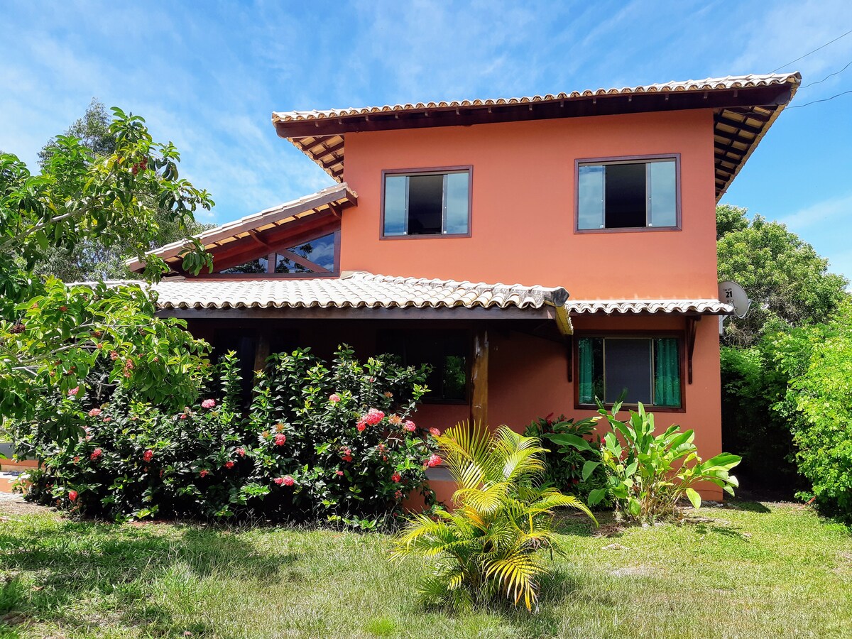 The exterior of a two-story rustic house is showcased, adorned with vibrant orange walls and a tiled roof. Lush greenery and flowering plants surround the entrance, contributing to the natural ambiance. Large windows allow light to enter, enhancing the connection with the outdoor landscape.