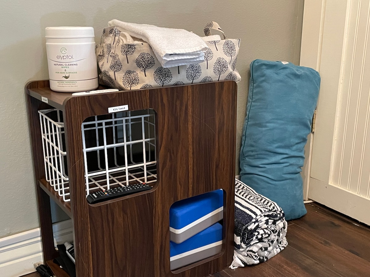 A wooden storage unit is positioned against a wall, featuring labeled compartments. Several neatly folded towels and a stack of blankets are stored alongside a blue cushion. A container labeled for personal care products sits atop the unit, enhancing the organized appearance.