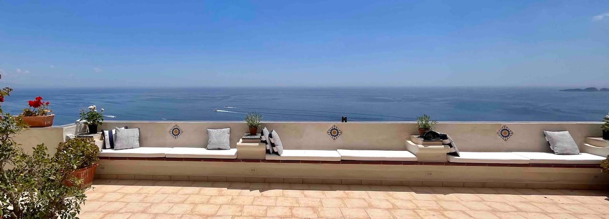 A spacious sun terrace overlooks the tranquil Tyrrhenian Sea, offering unobstructed coastal views. Soft seating with neutral cushions invites relaxation under a clear blue sky. Potted plants add a touch of greenery, while the tiled floor provides a warm, inviting space for outdoor enjoyment.