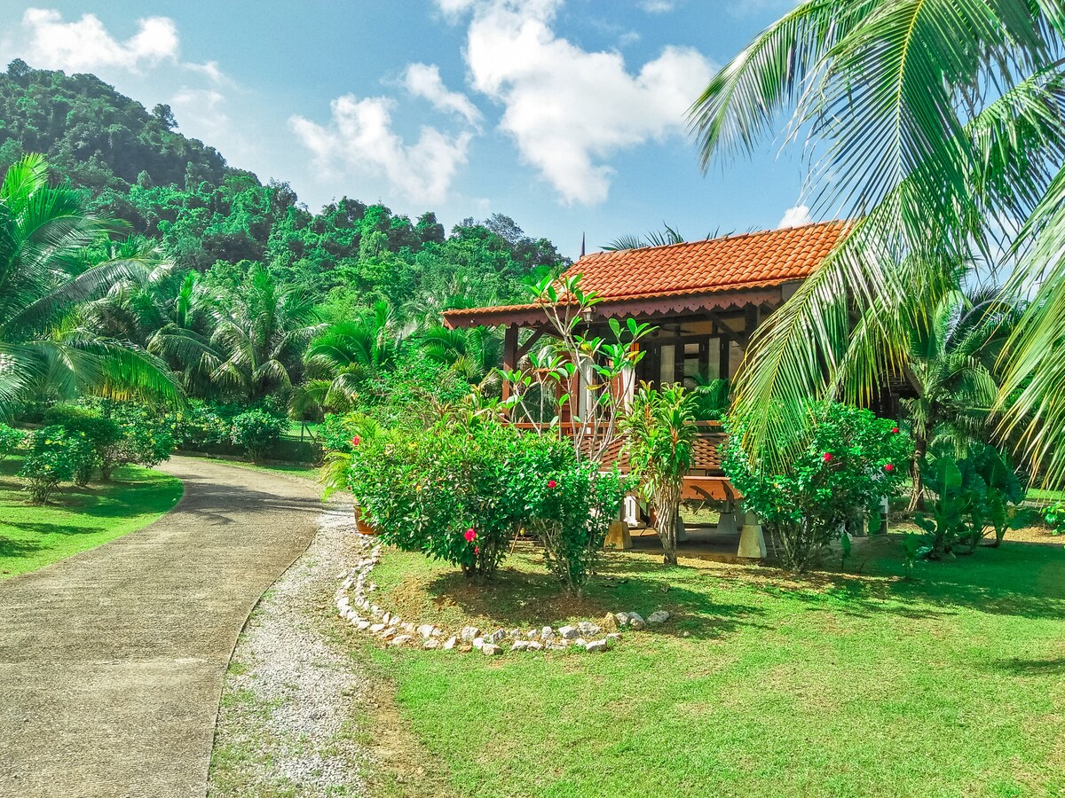 A charming villa is set against a backdrop of lush greenery and palm trees. A winding pathway framed by flowering plants leads to the wooden structure, which features a traditional Malay design and a sloped roof. Scenic hills provide an idyllic atmosphere.