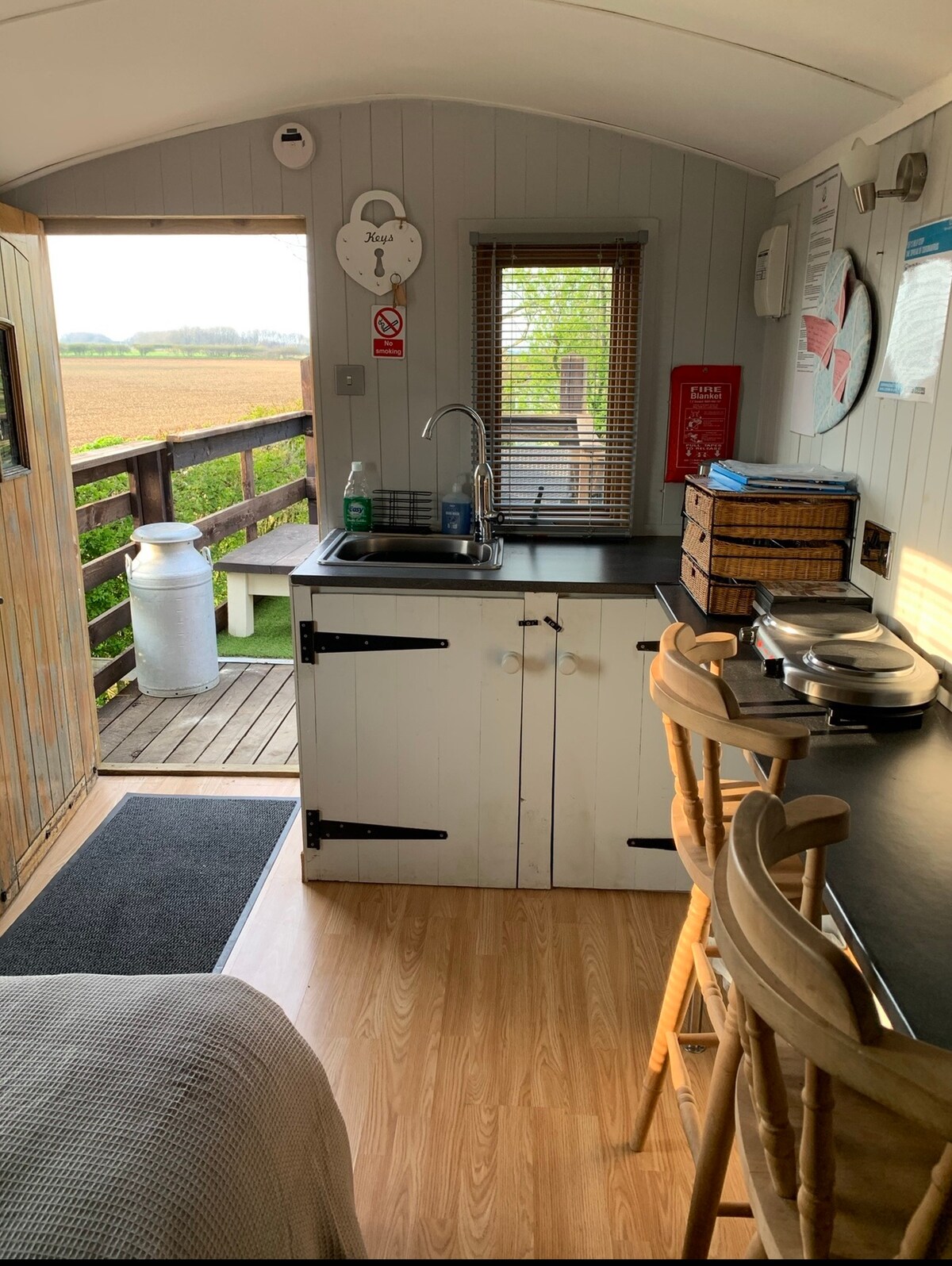 The interior of the shepherd's hut is presented, showcasing a compact kitchen area with a sink and countertop. Wooden bar stools are placed at a small table, while natural light filters through a window, revealing the rural landscape outside.