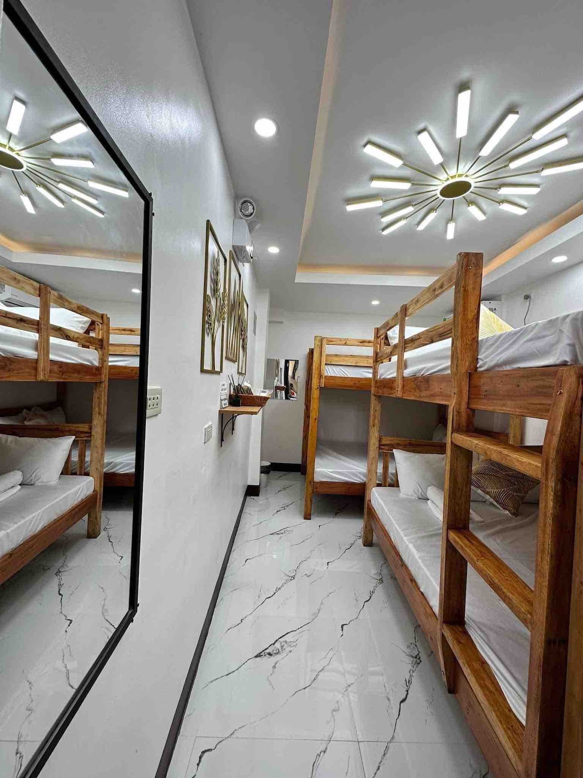 A well-lit bedroom features wooden double-deck beds against a neutral wall. An elegant ceiling fixture provides illumination, while a large mirror enhances the sense of space. The clean, white tiles add to the modern aesthetic, ensuring a comfortable and inviting environment.