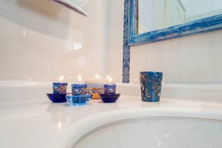 A serene bathroom setting is highlighted by an arrangement of blue glass candles placed on a white counter. Soft candlelight reflects off the surfaces, creating a calming ambiance. A decorative blue cup adds a touch of color, complemented by a matching blue-framed mirror in the background.