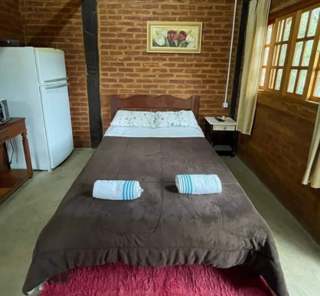 A cozy sleeping area features a double bed with a brown blanket and two rolled towels placed neatly on top. A simple bedside table sits beside the bed, and a wooden cabinet is visible in the corner. Natural light filters through the window, highlighting the brick wall.