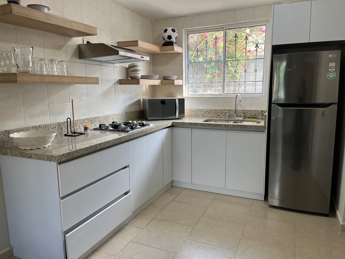 The kitchen features modern cabinetry with open shelves for dishware and decorative items. A stainless steel refrigerator stands against one wall, while a gas stove and microwave occupy the counter space. Natural light enters through a window adorned with floral views.