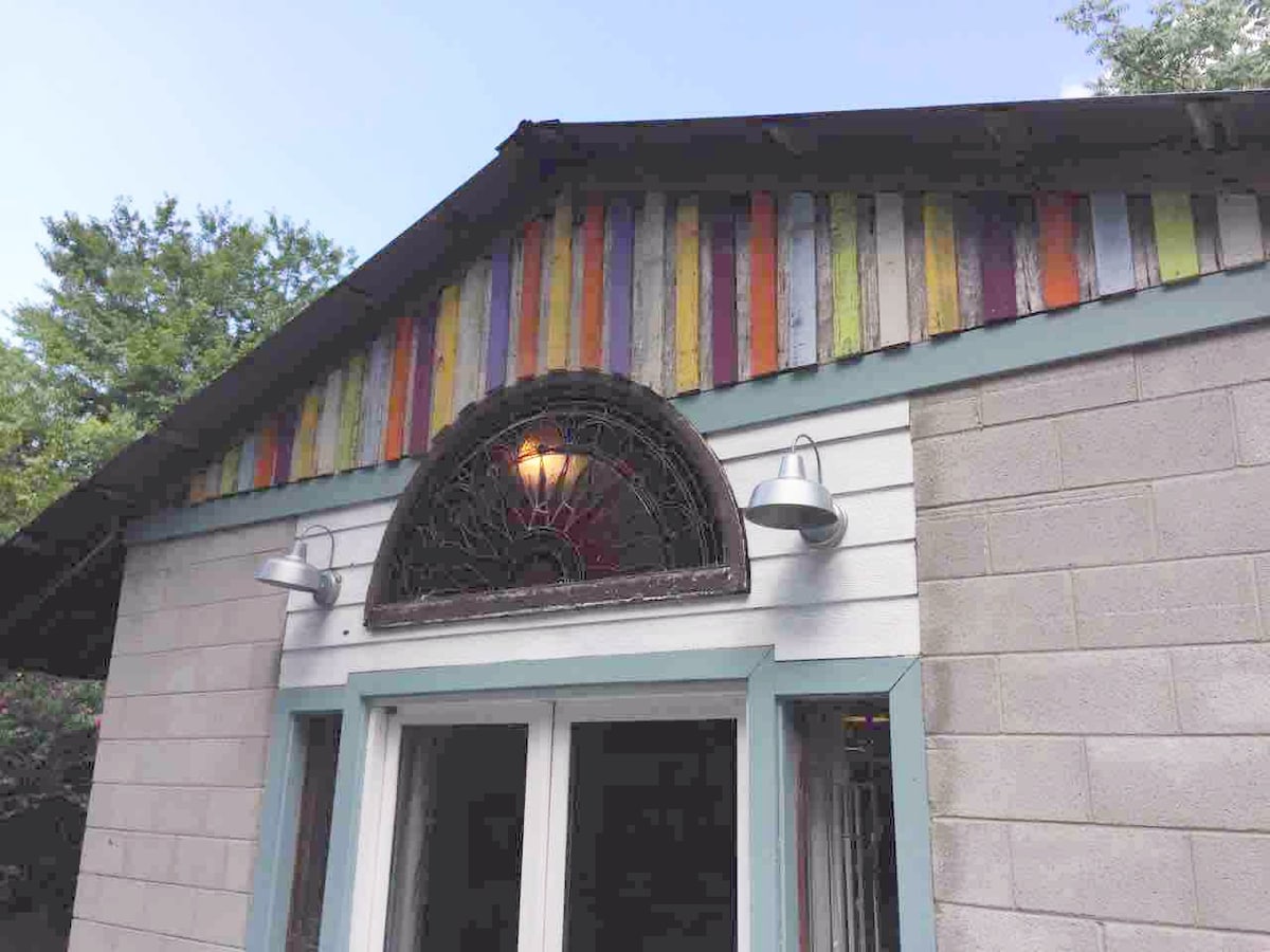The exterior of the studio showcases a distinctive architectural design, featuring a colorful wooden facade with vertical planks. An ornate, arched window is highlighted at the top, flanked by two stylish outdoor light fixtures, enhancing the inviting appearance of the entrance.