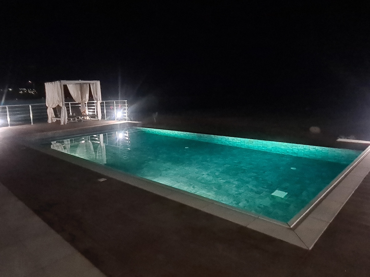 A large private pool is depicted, illuminated with a tranquil turquoise glow at night. Surrounding light fixtures enhance the pool area, while a shaded seating area with flowing fabric can be seen in the background.