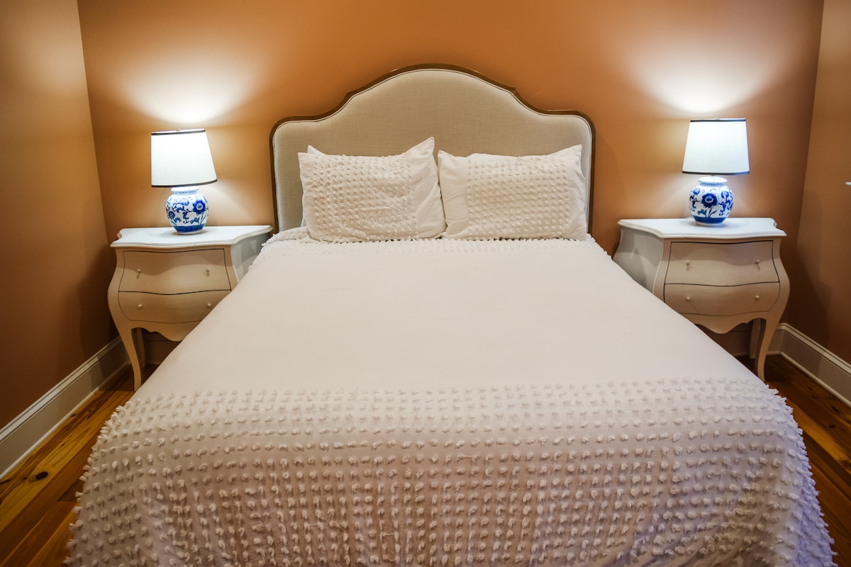A cozy sleeping area features a spacious bed dressed in a textured white covering. Nightstands with decorative lamps are positioned on either side, casting a soft glow. The walls are painted in a warm peach hue, enhancing the room's inviting feel.