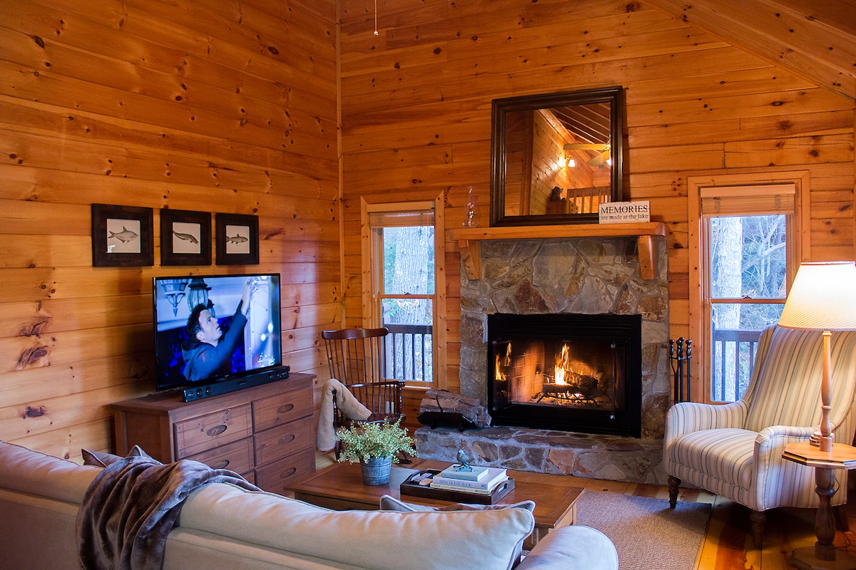 A cozy living area features a stone fireplace with a lit fire, surrounded by wooden walls. A flat-screen TV is positioned on a wooden cabinet, and a plush sofa with a throw blanket invites relaxation. Natural light enters through nearby windows.