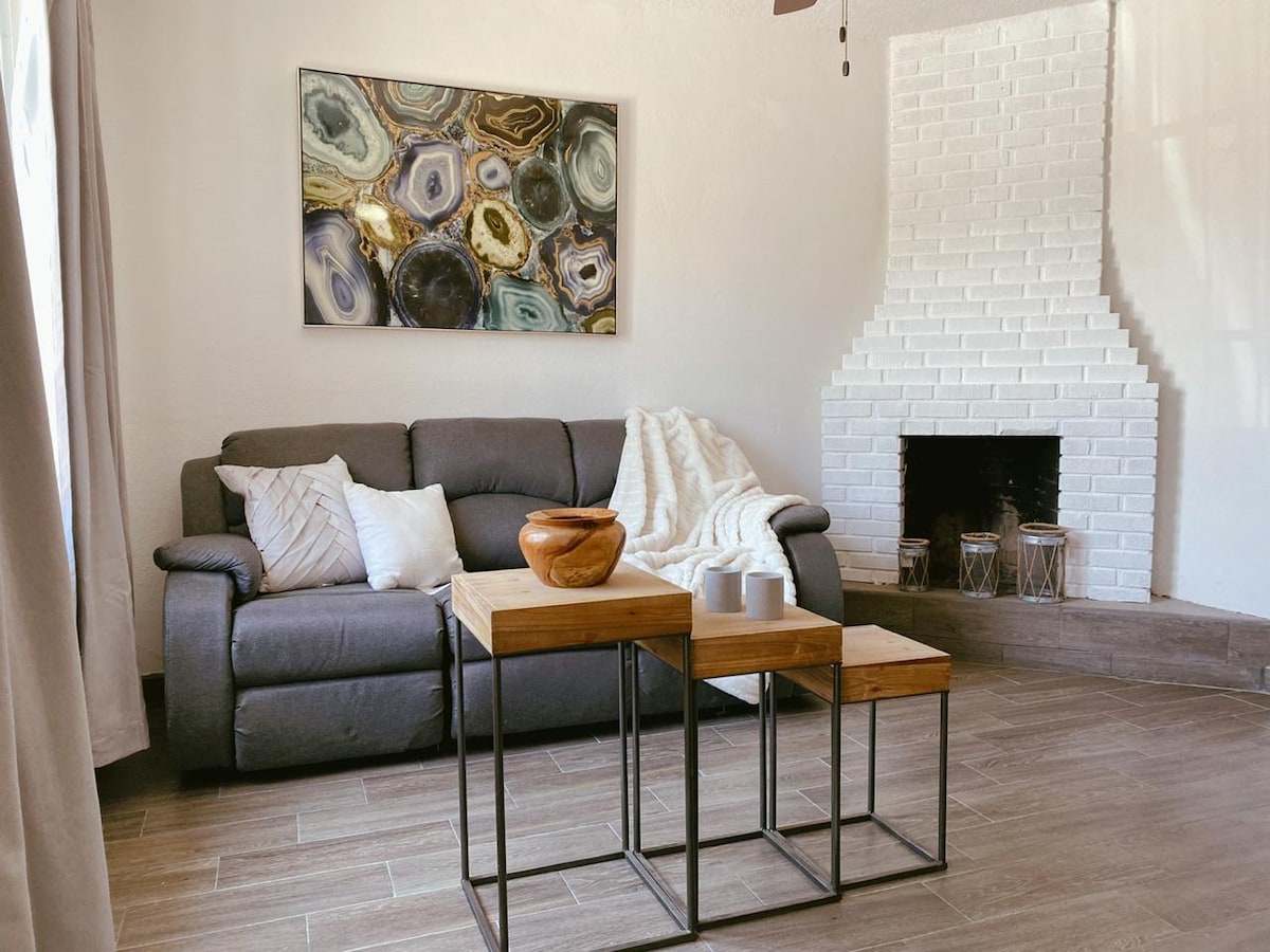 A living space features a comfortable gray sofa, layered with a soft throw blanket. Two wooden side tables with metal legs are positioned in front. A decorative bowl and mugs rest on one table, while a white brick fireplace serves as a focal point, complemented by wall art.