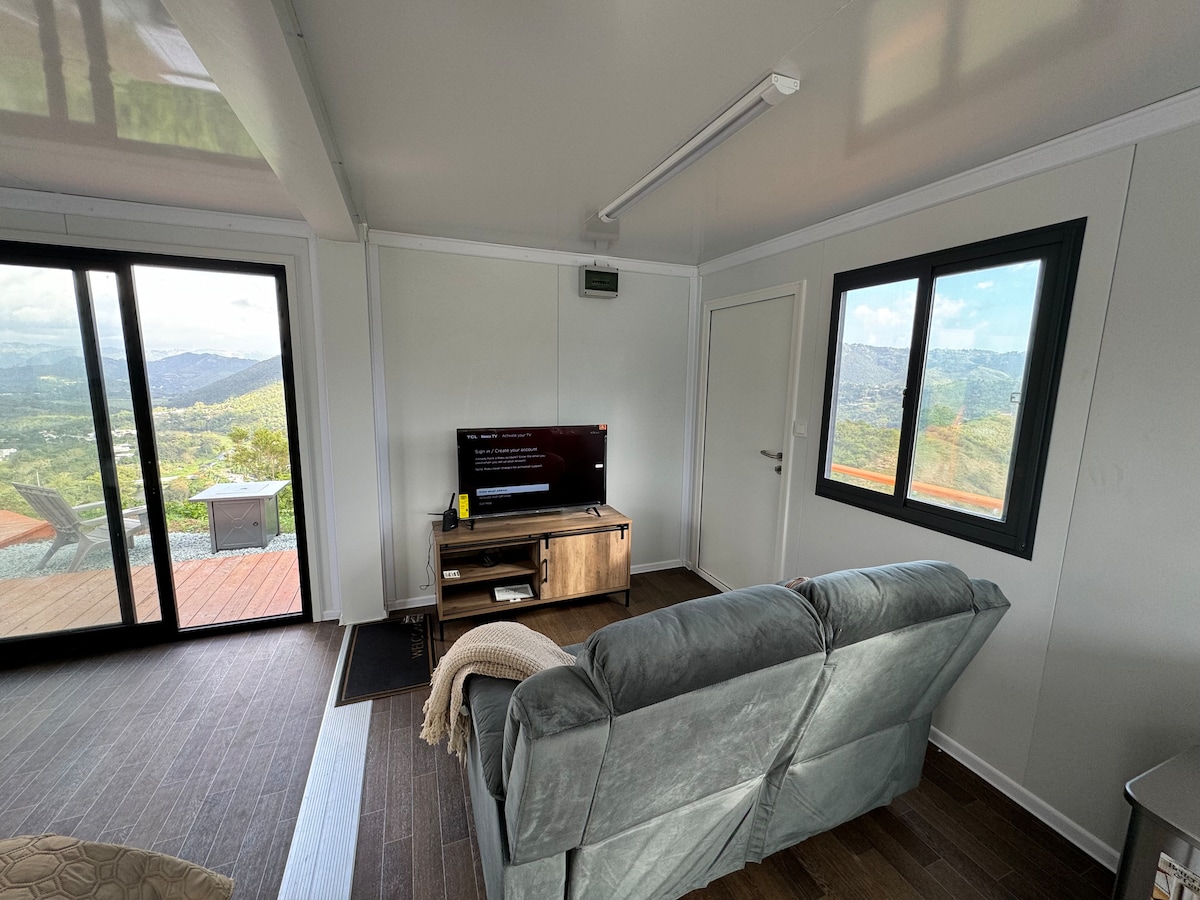 A cozy living area features a comfortable grey sofa facing a television on a wooden stand. Large windows provide expansive views of the surrounding mountains and landscape. Natural light brightens the space, complemented by minimalist decor.