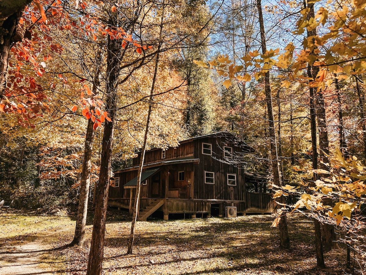 A rustic cabin is nestled among vibrant autumn foliage, with leaves in shades of orange and yellow. Sunlight filters through the trees, illuminating the wood exterior and creating a tranquil scene. A pathway leads to the entrance, surrounded by a peaceful natural setting.