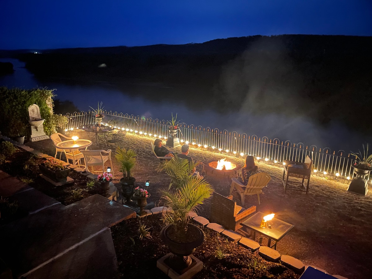 An outdoor seating area is illuminated at night, featuring several chairs around a fire pit. Soft lighting outlines the perimeter, while the view of a river and rolling hills is visible in the background, enhancing the tranquil ambiance.