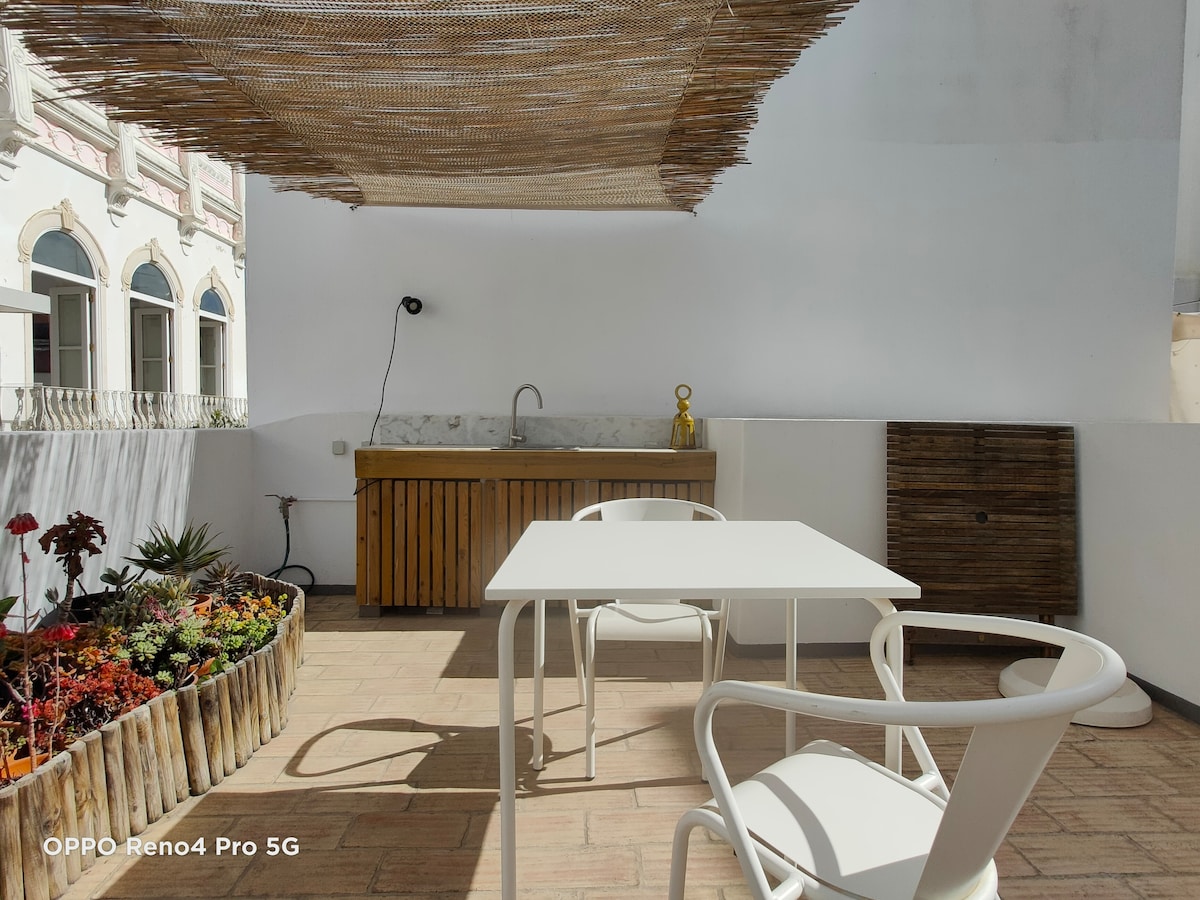 An outdoor terrace features a wooden kitchen area with a sink and ample counter space. A simple white table and chairs are arranged nearby. The space is shaded by a natural woven canopy, with a circular planter filled with vibrant plants adding color.