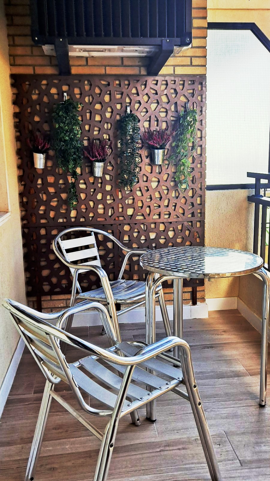 A small outdoor balcony features two metal chairs and a glass-top table. A decorative wooden panel displays hanging plants, enhancing the space's natural feel. The balcony is bathed in soft natural light, creating a serene spot for relaxation.