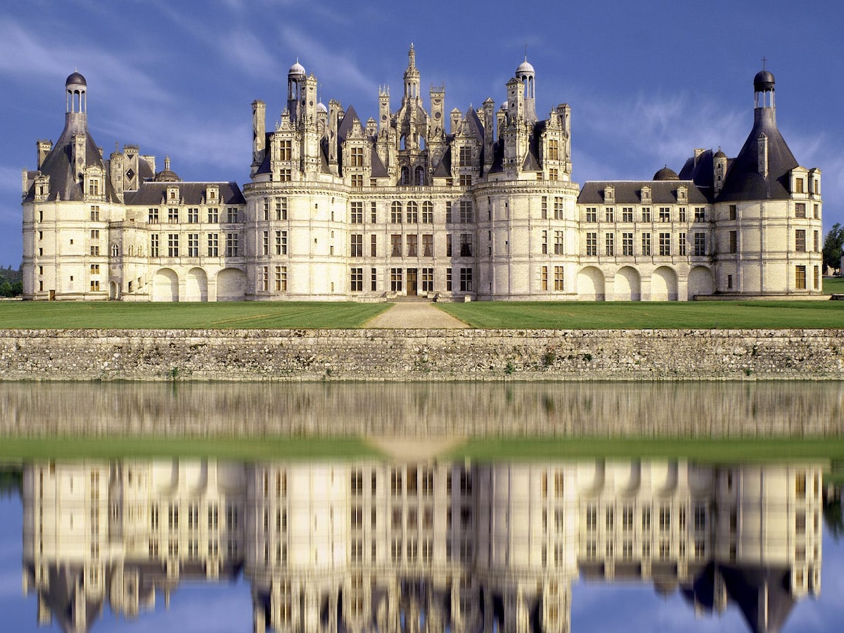 A grand chateau is reflected in calm waters, showcasing intricate architecture with multiple towers and sculptural details. The stone facade is complemented by expansive lawns, providing a serene and historical setting.