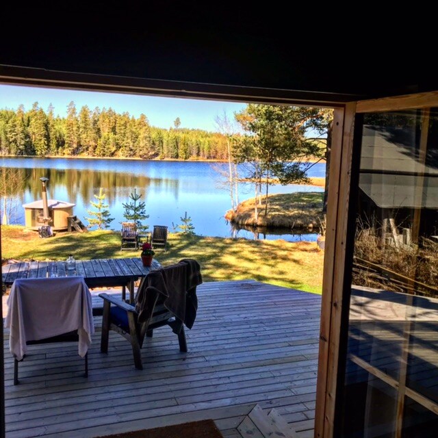 A tranquil lakeside view is framed by the open doorway of a rustic cabin. A spacious wooden deck features a table and chairs, inviting relaxation. The calm water reflects the surrounding trees, enhancing the serene landscape. Sunlight filters through the vibrant green foliage.