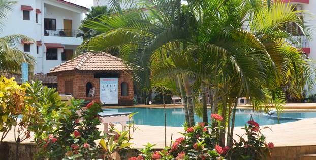 A serene swimming pool is surrounded by lush greenery and colorful flowering plants. A palm tree adds a tropical touch, while a thatched-roof gazebo is visible in the background, enhancing the relaxing outdoor environment.