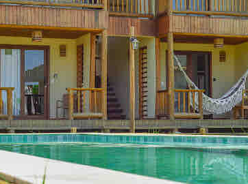 A refreshing swimming pool is bordered by a stone deck and wooden railing. Wooden balconies are visible on the second floor, offering access to outdoor spaces. A light-colored hammock hangs between two posts, creating a relaxing spot near the pool area.