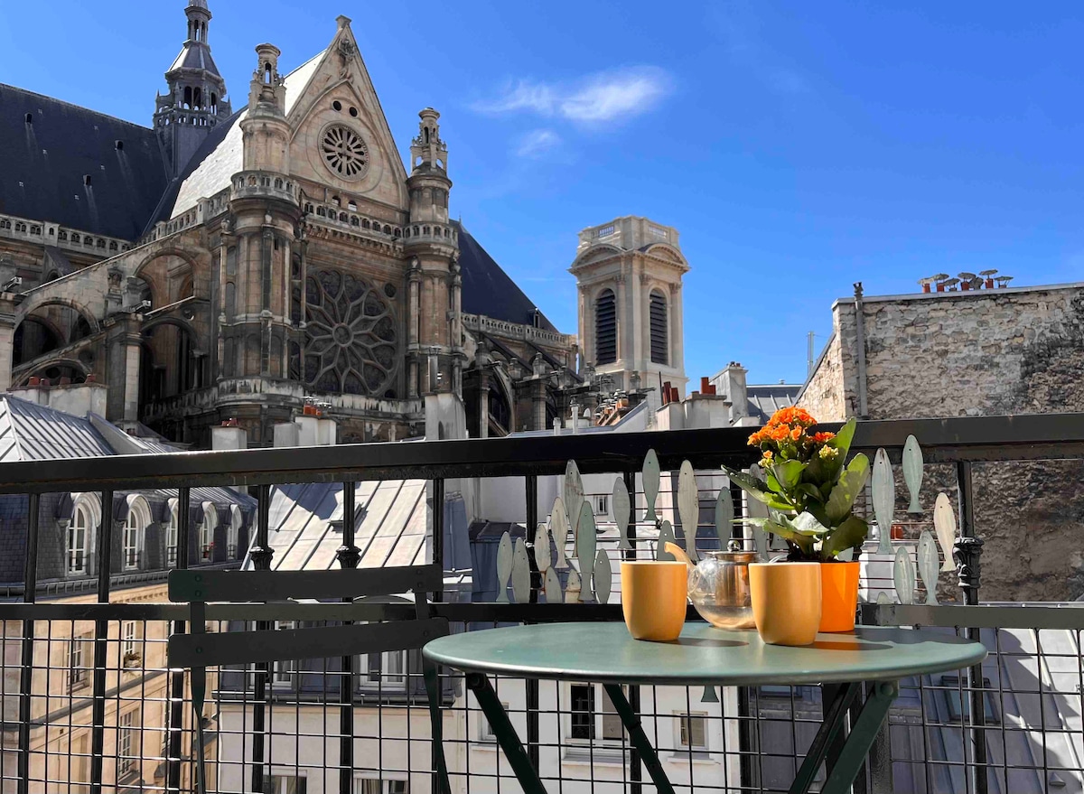 The balcony features a small green table accompanied by two cups and a glass. A floral arrangement adds a touch of greenery. In the background, the intricate architecture of the Basilica of Saint-Eustache is highlighted against a clear blue sky.