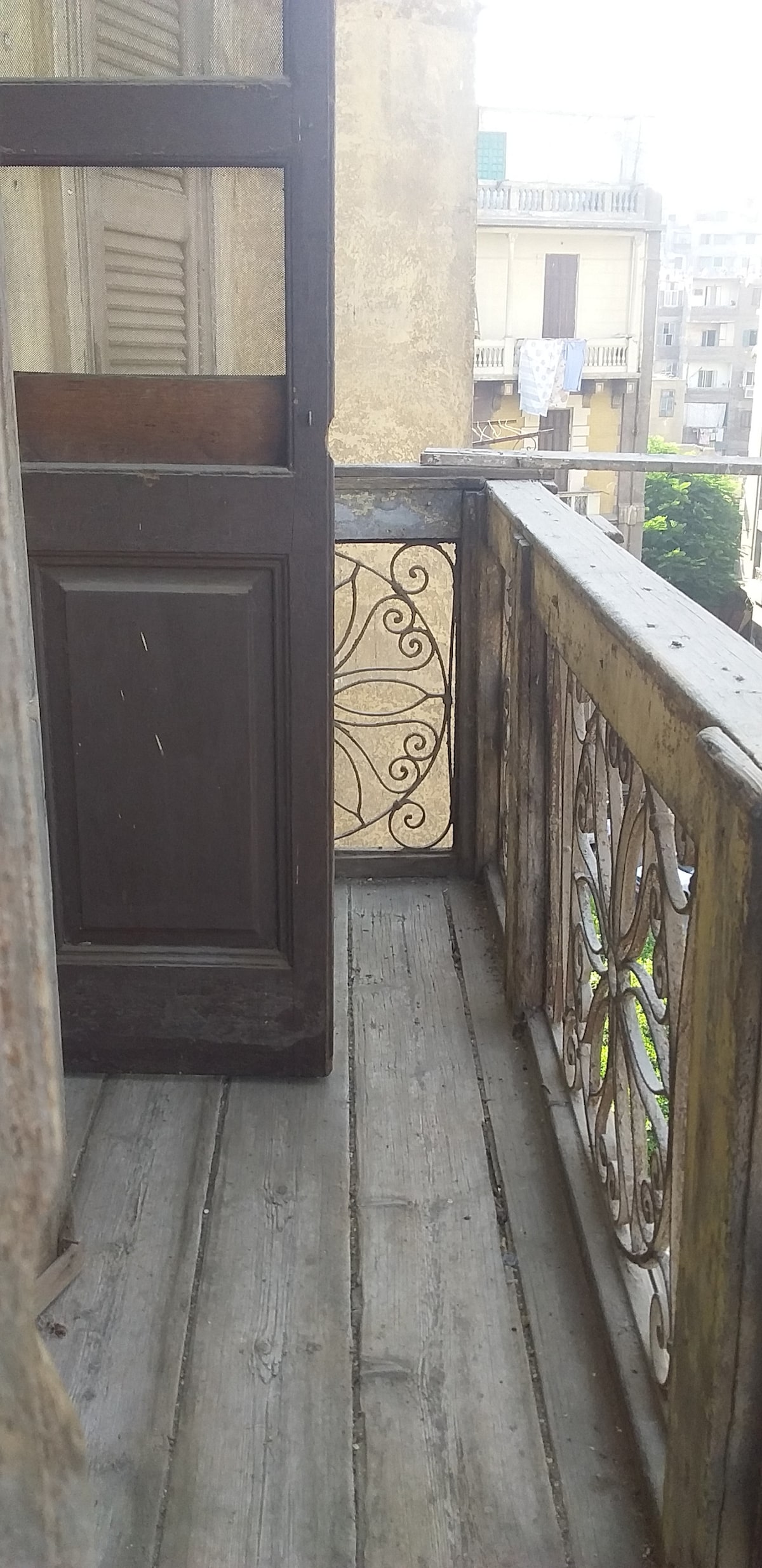 An exterior balcony is visible, featuring ornate metal railings and rustic wooden flooring. The door to the balcony is partially open, allowing a glimpse of the surrounding architecture and greenery just outside.
