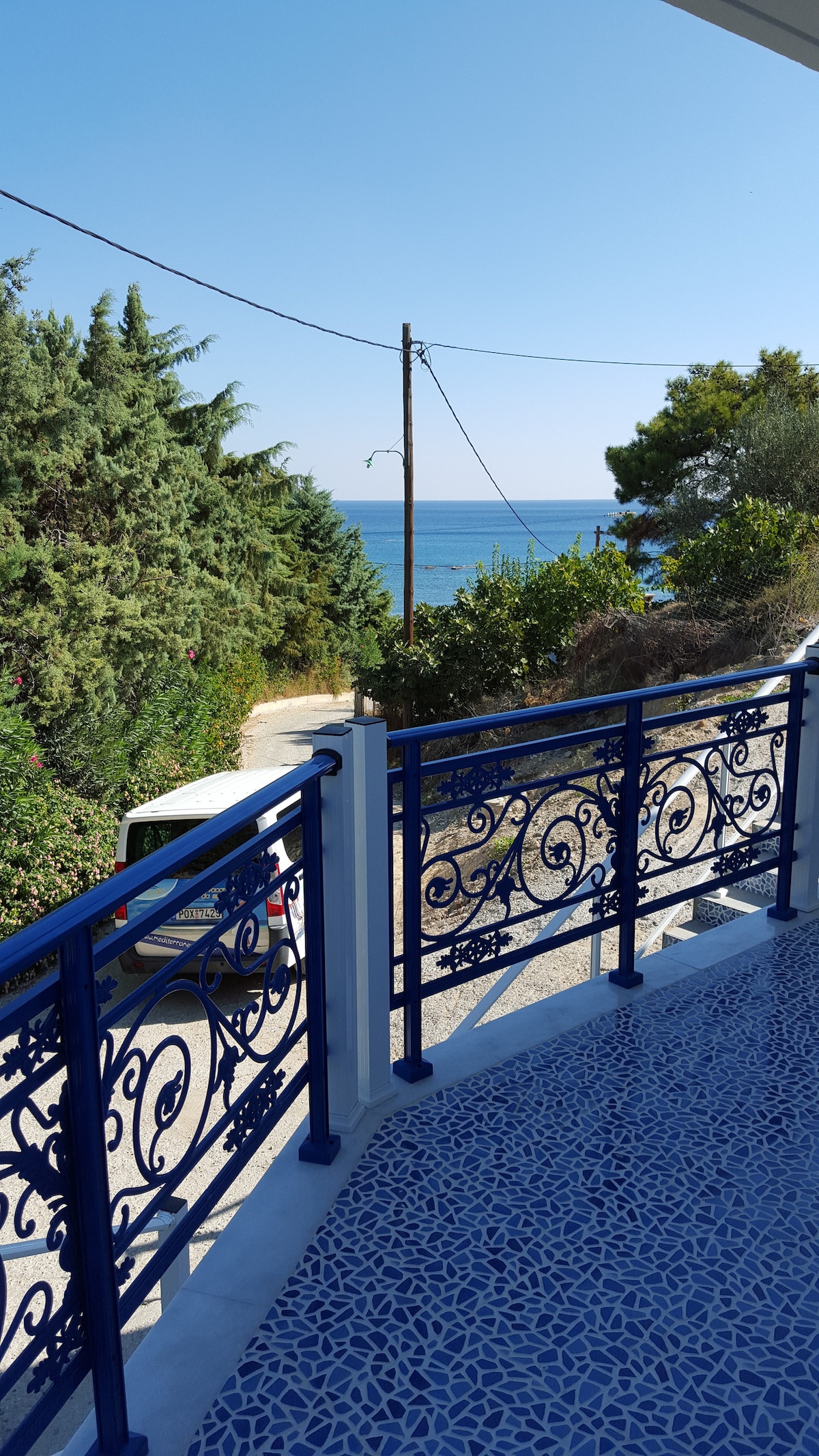 A balcony view reveals a calm sea in the distance, framed by lush greenery. The balcony railing features intricate blue ironwork, contrasting against the light-colored flooring. The sky is clear, showcasing a bright atmosphere.