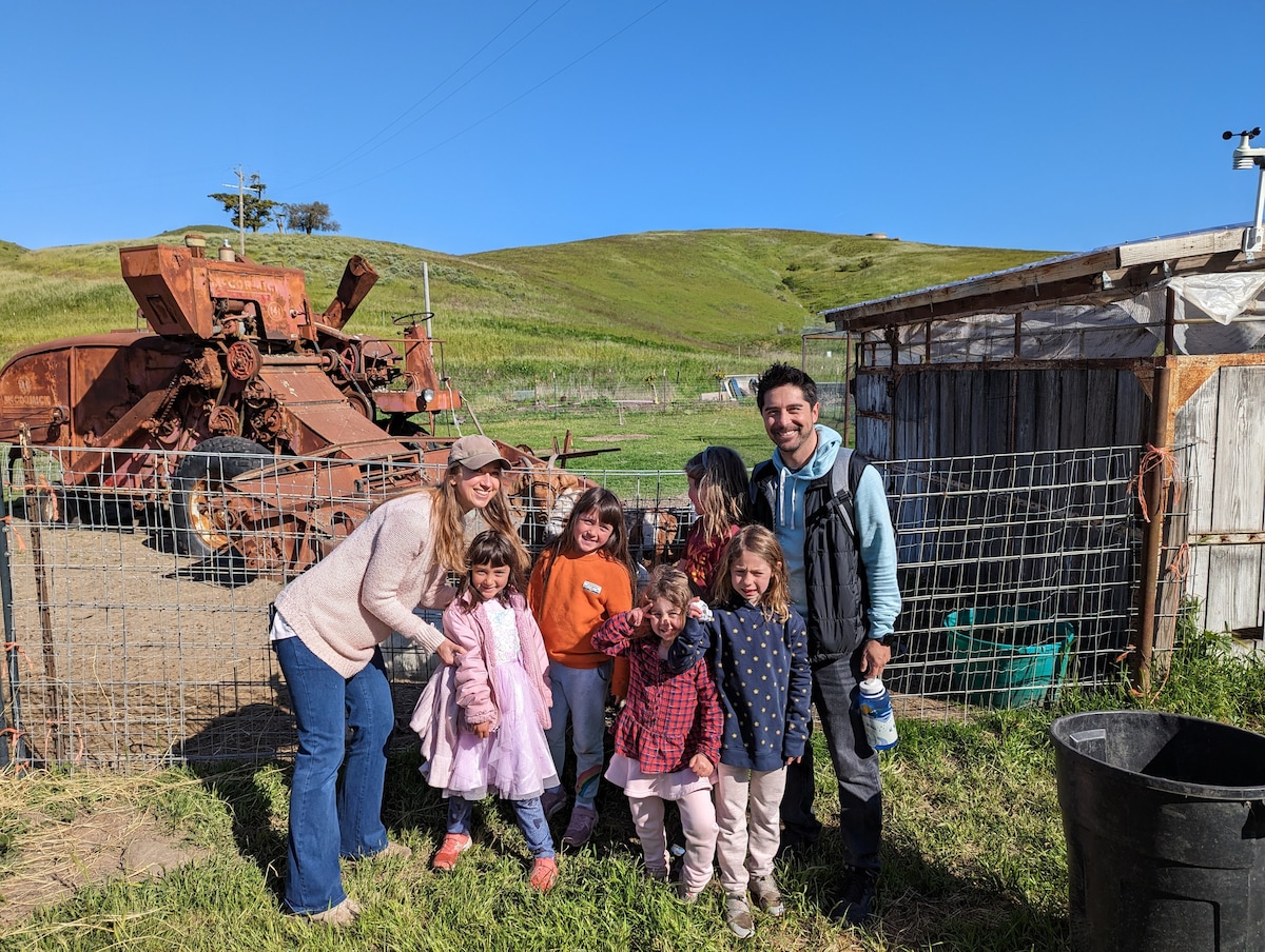 A group of seven individuals is gathered near a vintage farming vehicle, set against a backdrop of rolling green hills. The area appears rural, with a weathered shed visible nearby. Bright sunlight enhances the outdoor scene, contributing to a lively atmosphere.