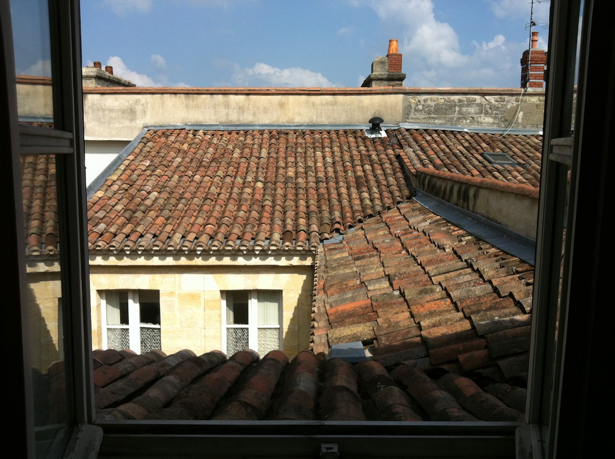 A view is captured through a window, showcasing rooftops with traditional terracotta tiles. The serene sky is punctuated with fluffy clouds, enhancing the historical character of the surrounding buildings and architecture.