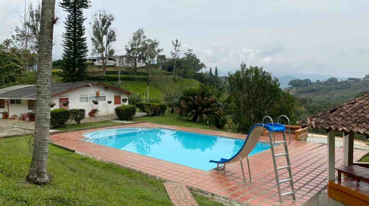 A sparkling swimming pool is surrounded by well-maintained green grass and tropical plants. A slide is positioned at one end, with a ladder nearby. The landscape is accented by gently rolling hills and a distant mountain backdrop, creating a serene outdoor setting.