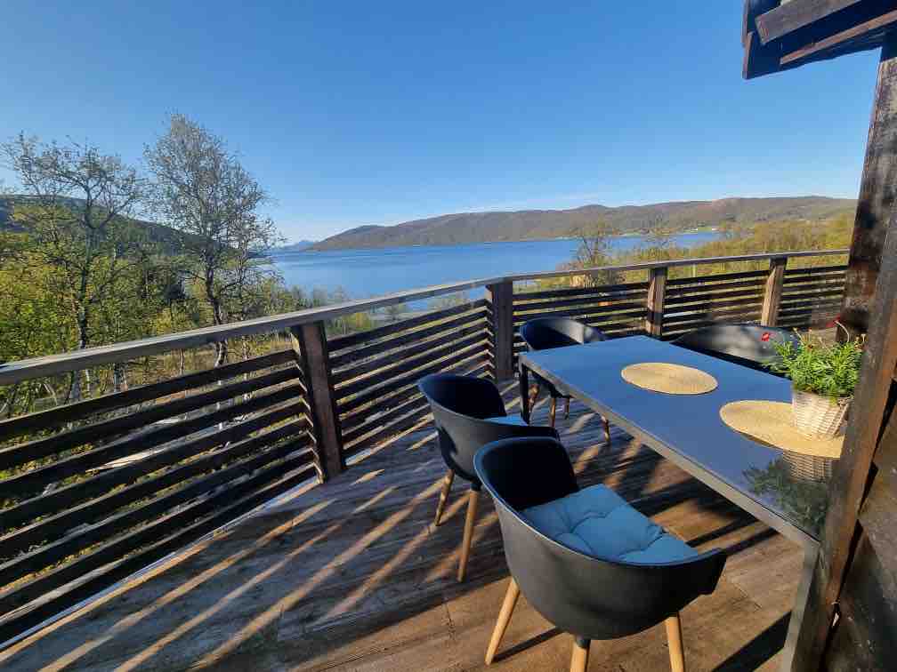 An outdoor terrace features a modern table set for dining, surrounded by black chairs. The view overlooks calm waters and distant hills, framed by lush greenery and clear blue skies, creating a serene space for relaxation.