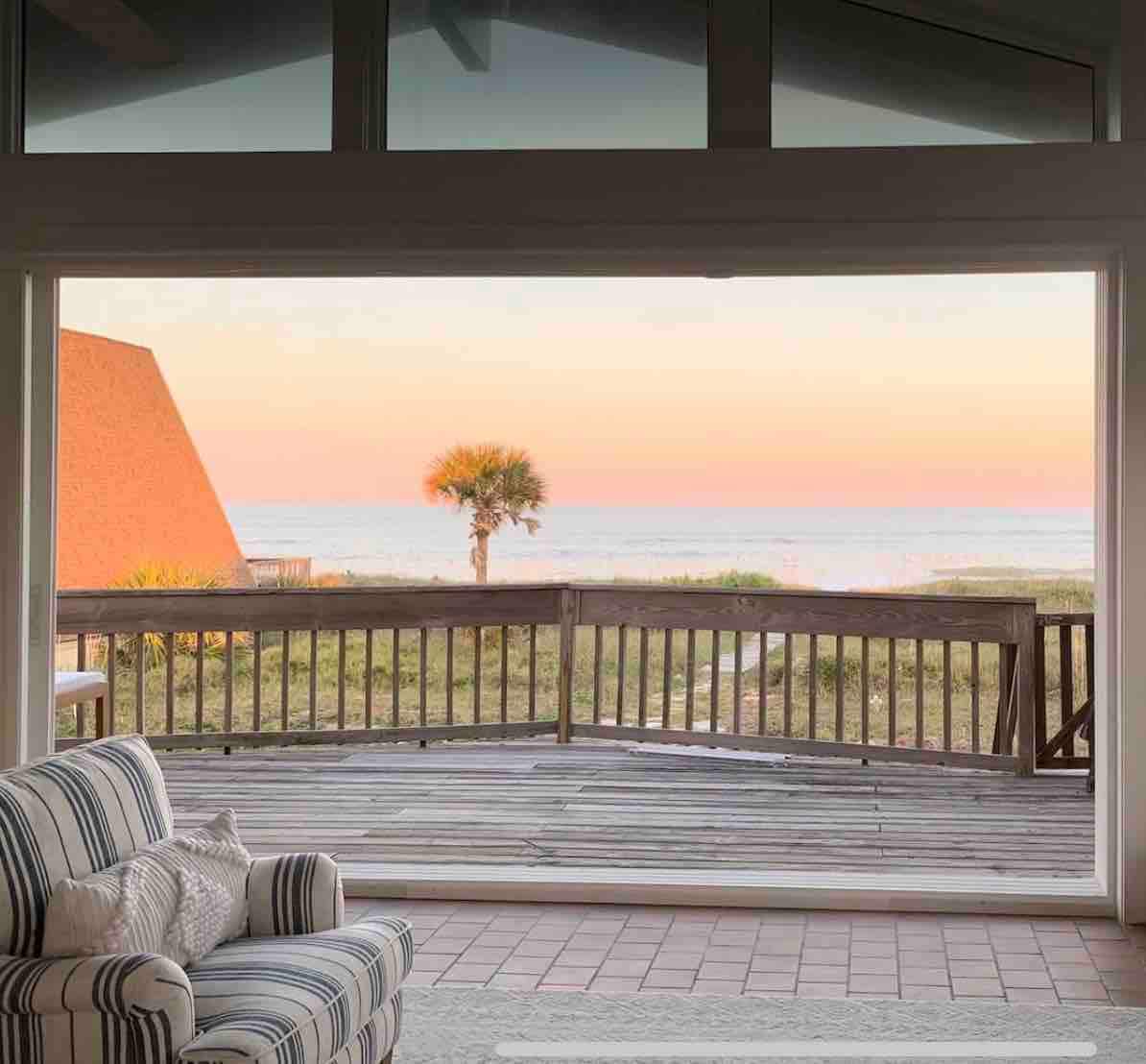 A spacious living area features large sliding glass doors framing stunning ocean views. The scene includes a wooden deck and a solitary palm tree against a soft pastel sunset sky.
