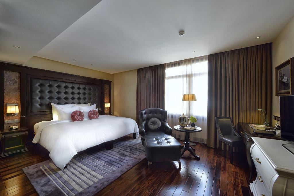 A spacious bedroom is presented with a large bed featuring a tufted headboard. A comfortable armchair and small side table are positioned next to a window dressed in thick curtains. Warm wood flooring enhances the inviting ambiance of the room.