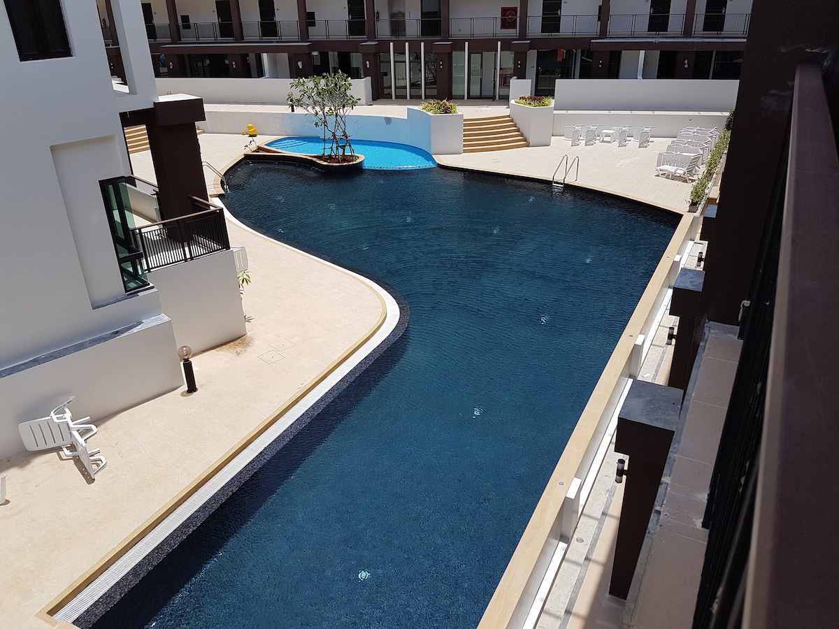 A curved outdoor swimming pool is visible, surrounded by lush greenery and outdoor lounge chairs. Steps lead down to the pool, while a children's area with shallow water is situated nearby. The setting is spacious and invites relaxation under clear skies.