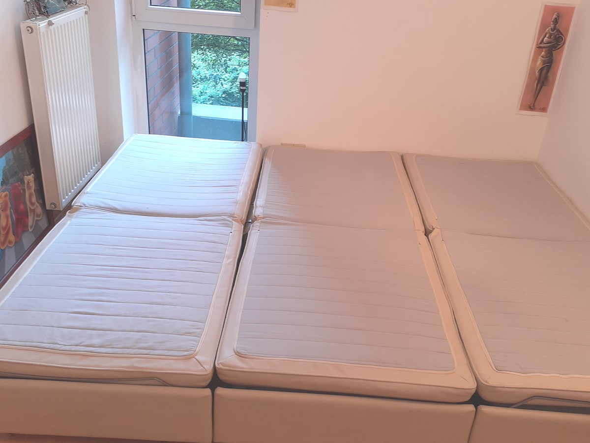 A simple sleeping arrangement is presented with multiple foam mattresses, neatly arranged on the floor. Natural light enters through a nearby window, illuminating the space. The room features a minimalistic decor with a small radiator visible near the entrance.