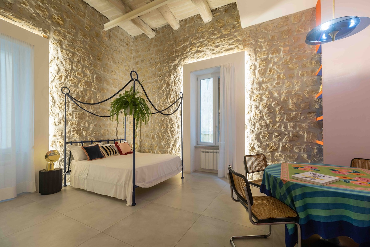 A room with natural stone walls features a spacious bed adorned with decorative pillows. A small dining table with colorful tablecloth and woven chairs complements the space. Soft light filters through sheer curtains, highlighting the room's unique texture.