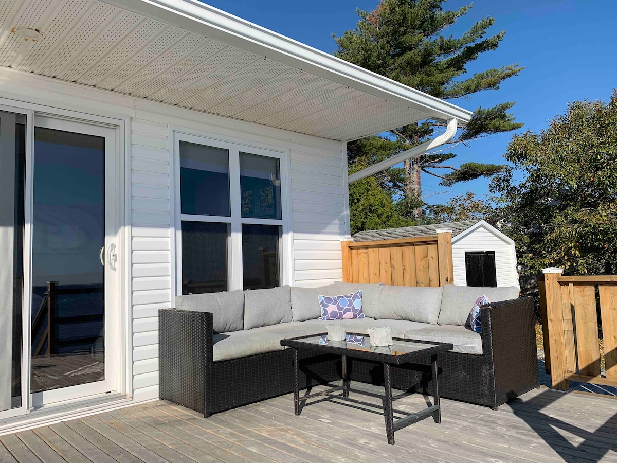 A spacious outdoor deck features a comfortable sectional seating area with light-colored cushions. A glass-topped coffee table is positioned in front, surrounded by wooden railings. Green trees provide a backdrop against the clear blue sky, creating a serene outdoor setting.