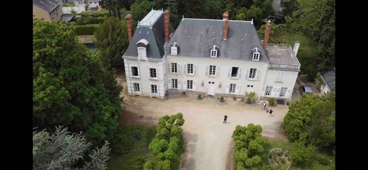 An aerial view of a grand 18th-century house is showcased, framed by lush greenery. The light-colored façade features multiple windows and a steeply pitched roof, with brick chimneys rising above. The spacious driveway is lined with symmetrical hedges leading to the entrance.