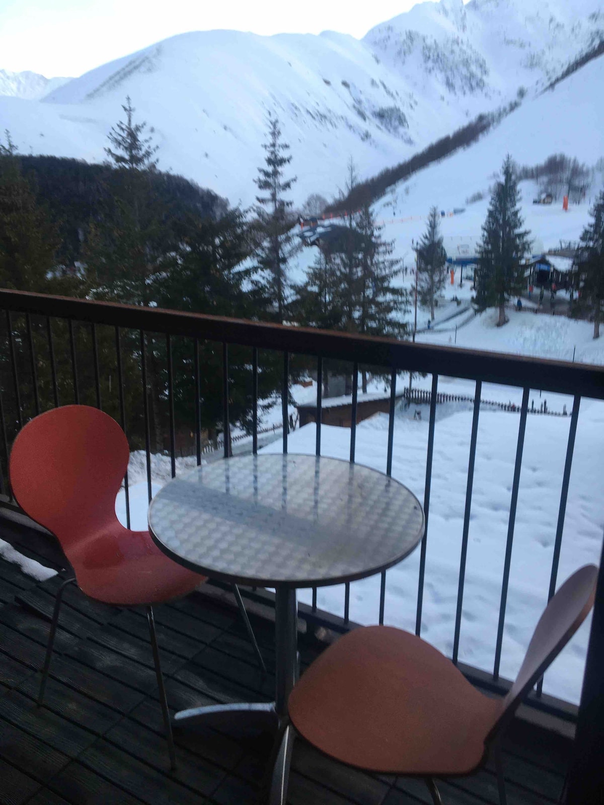 A balcony features a round glass table and two orange chairs, overlooking a snowy landscape. Pine trees dot the view, while distant ski slopes are visible, creating a serene mountain setting.