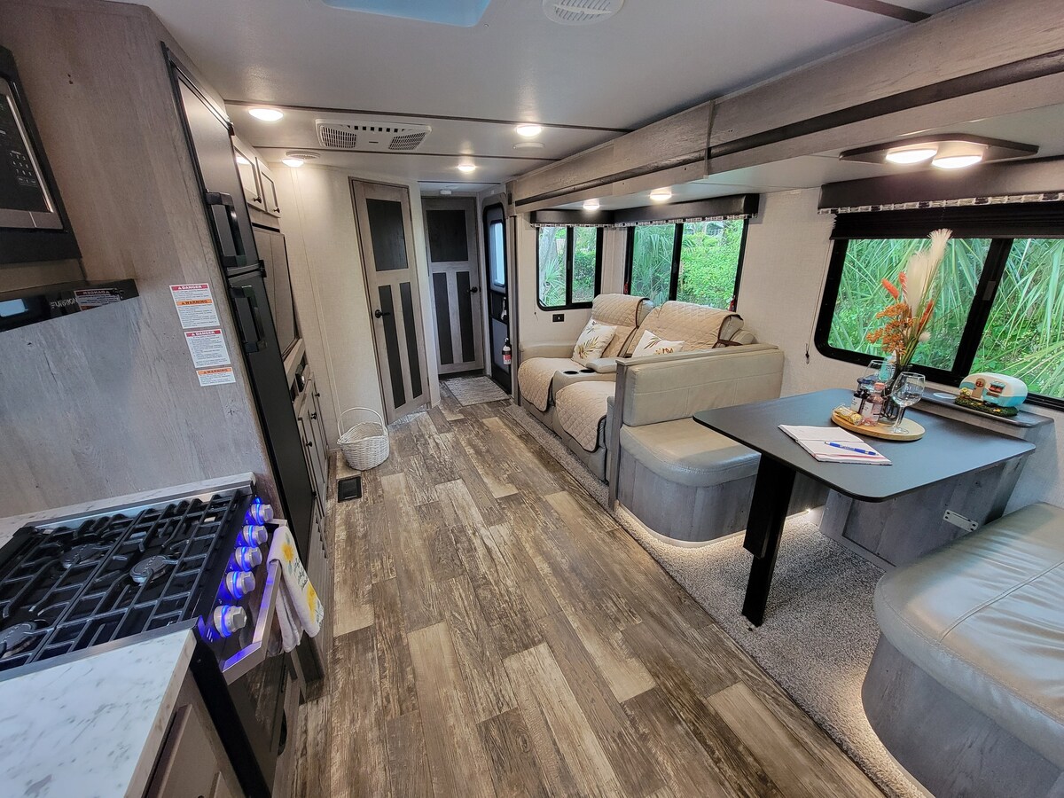 The interior of the RV showcases a spacious layout with a well-equipped kitchen on the left and a comfortable seating area on the right. Natural light enters through multiple windows, and a neutral color palette creates a calming environment. Stylish decor includes a dining table and cozy sofa.