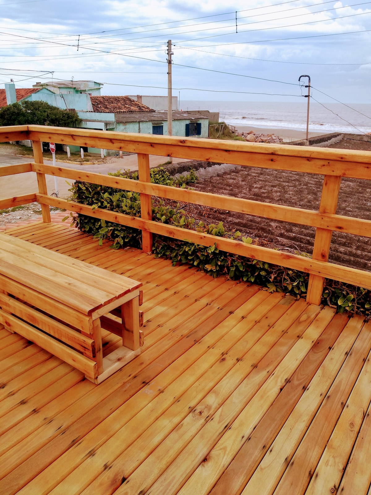 A wooden deck is featured with a simple bench, providing a clear view of the ocean in the distance. The surrounding railing is complemented by greenery, enhancing the outdoor space's connection to nature.