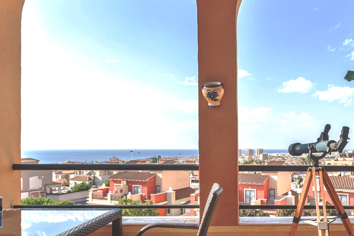 A balcony view features a telescope positioned beside a wicker chair, overlooking a vibrant cityscape that extends towards the sea. The clear sky provides a bright atmosphere, while decorative elements, including a wall-mounted pottery piece, add character to the space.