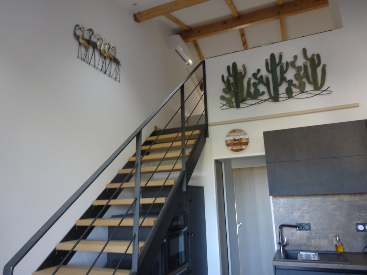 An open space showcases a stylish staircase leading to a mezzanine, framed by wooden beams. Wall decor includes a metal sculpture and cacti artwork, adding character. The modern kitchen area features sleek cabinetry and appliances, enhancing functionality and aesthetic appeal.