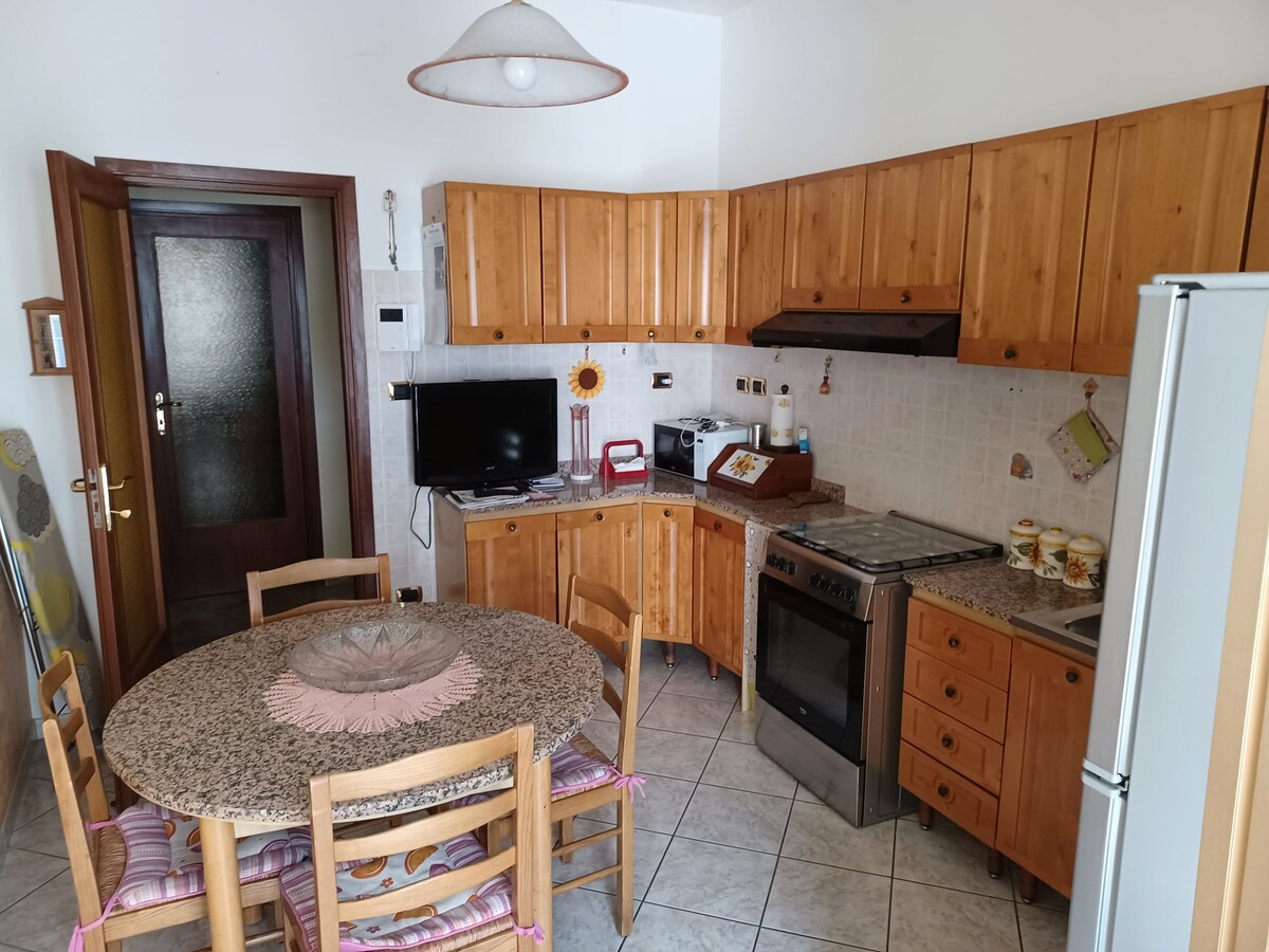 The kitchen features wooden cabinetry and a round dining table surrounded by six wooden chairs. A gas stove and oven are positioned alongside an ample countertop. A television is mounted on the wall, and light enters through the door leading to the entrance.