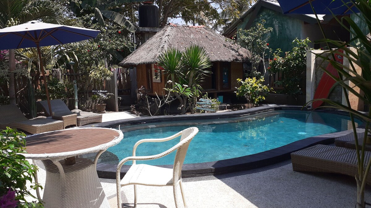 A refreshing swimming pool is surrounded by lounge chairs and umbrellas, providing shaded seating. Tropical plants and trees are visible around the pool area, while a thatched-roof structure adds a charming element in the backdrop.