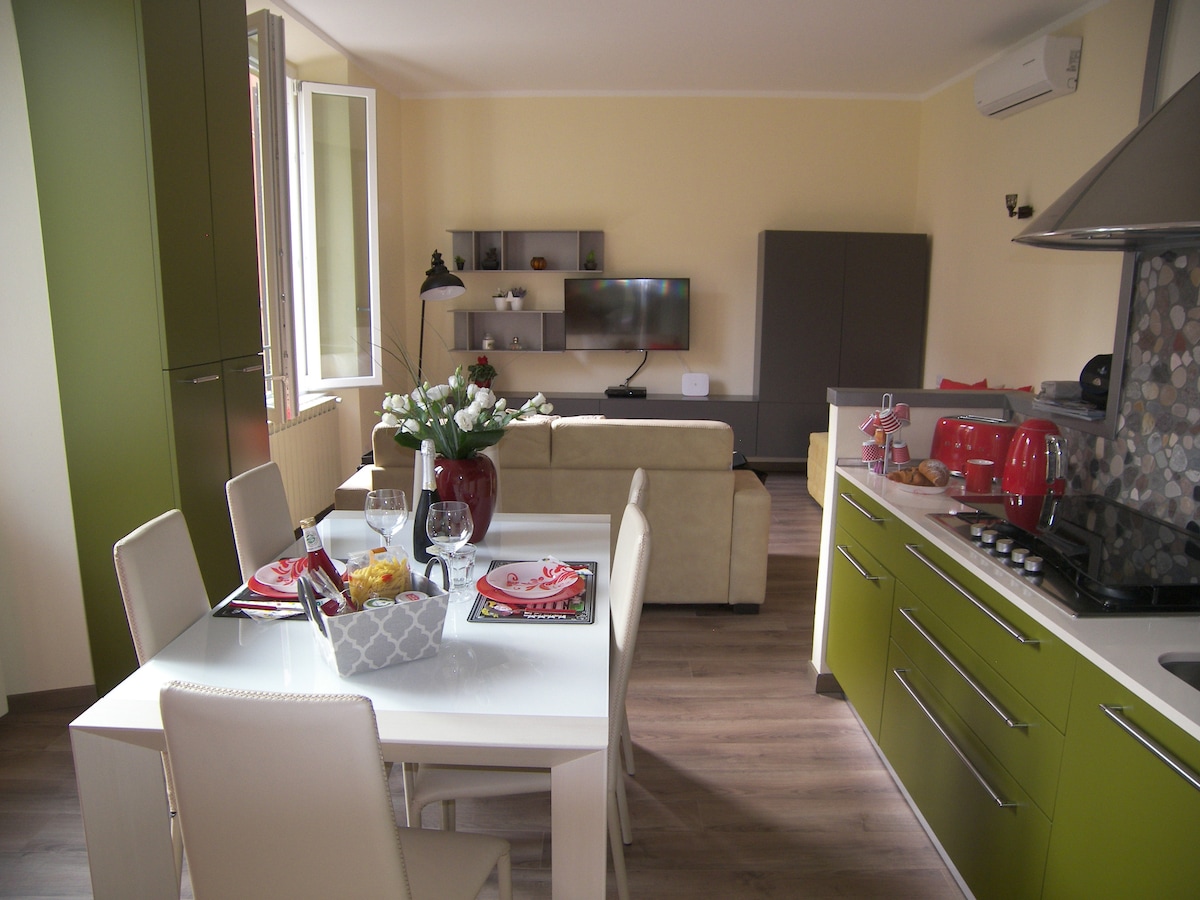 A modern kitchen and dining area features a white table set for four, including glassware and colorful plates. Green cabinets add a vibrant touch. In the background, a living space with a light-colored sofa and wall-mounted TV is visible, enhancing the open-plan design.