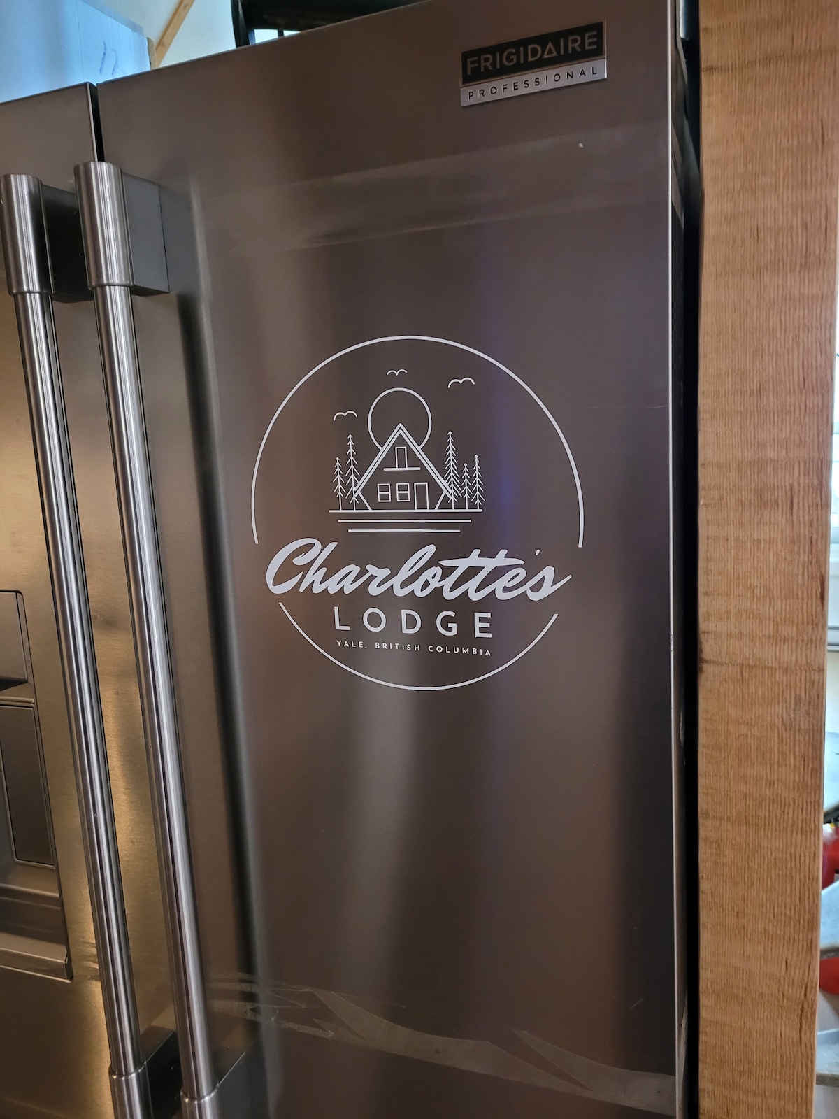 A modern refrigerator with a sleek stainless steel finish is prominently displayed. The Charlotte's Lodge logo is elegantly featured on the front, showcasing the name and location of the lodge in Yale, British Columbia.