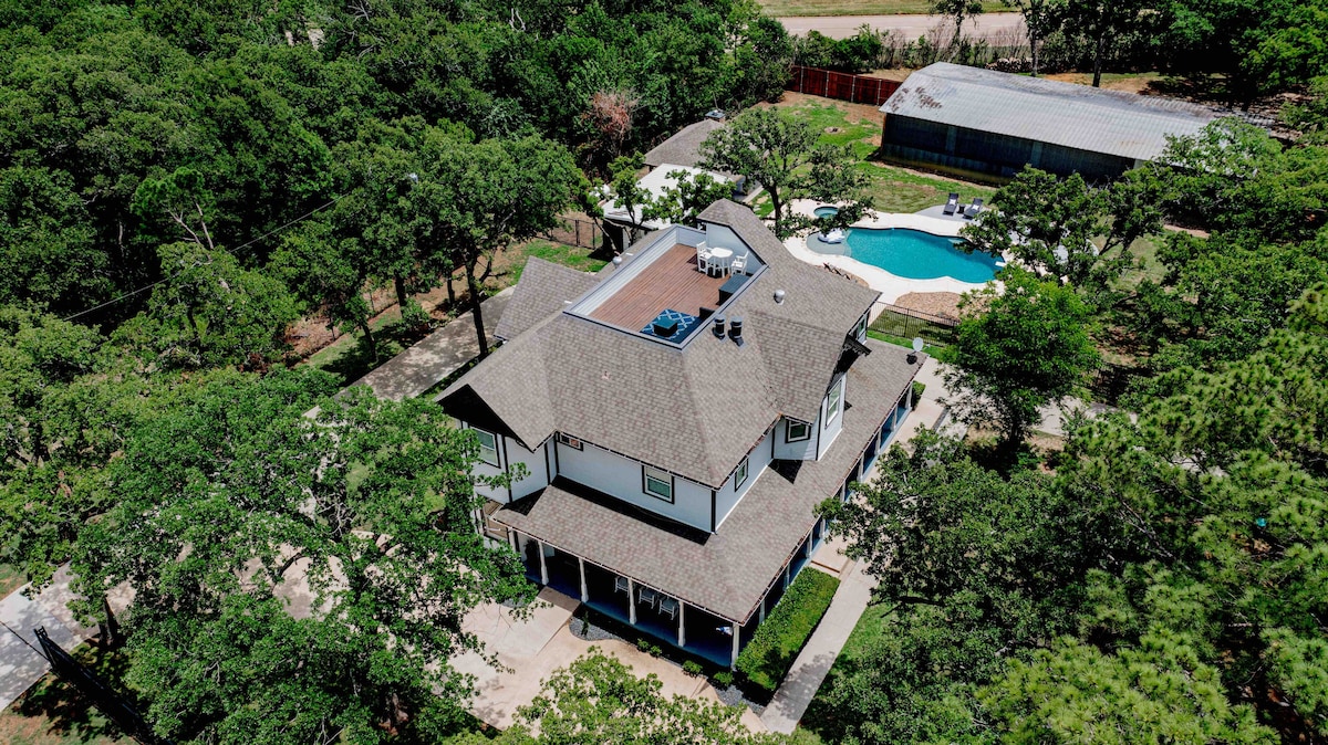 An aerial view showcases a large Victorian home surrounded by lush greenery. The spacious rooftop deck is visible, as well as a sizable pool in the backyard, bordered by lounge chairs and shaded areas. The property offers a tranquil natural setting.