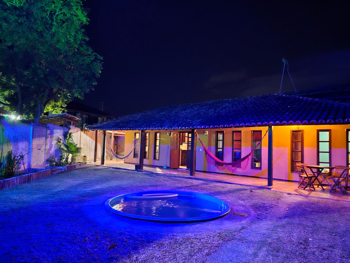 The outdoor area is illuminated at night, featuring a circular pool surrounded by vibrant lighting. Hammocks are hung between trees, and the house is visible in the background, showcasing windows and a welcoming entryway. Lush grass completes the tranquil setting.