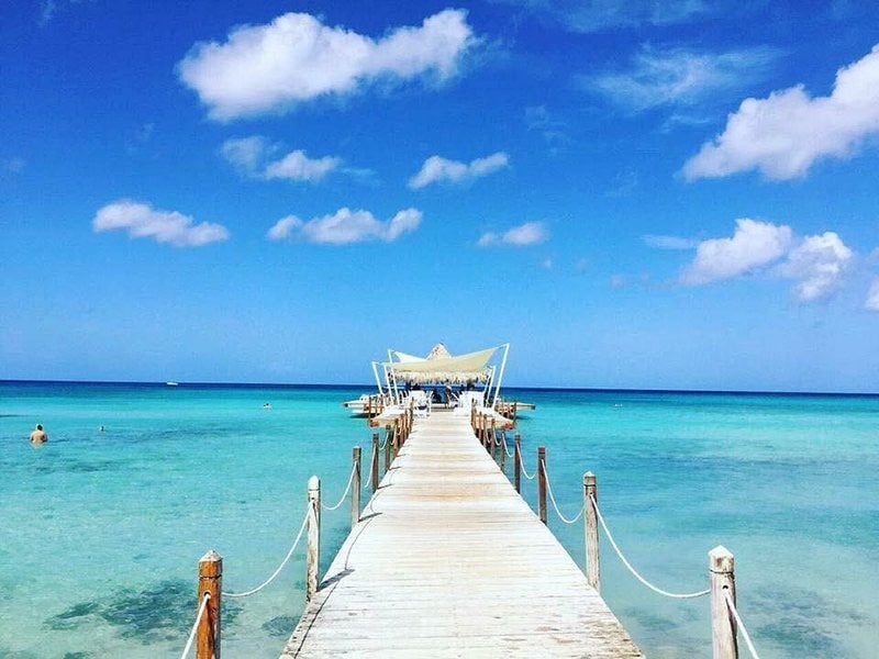 A long wooden pier extends over calm turquoise waters, leading to a shaded area with a canopy. Light clouds float across a bright blue sky, while a few swimmers can be seen in the distance, enjoying the clear water.
