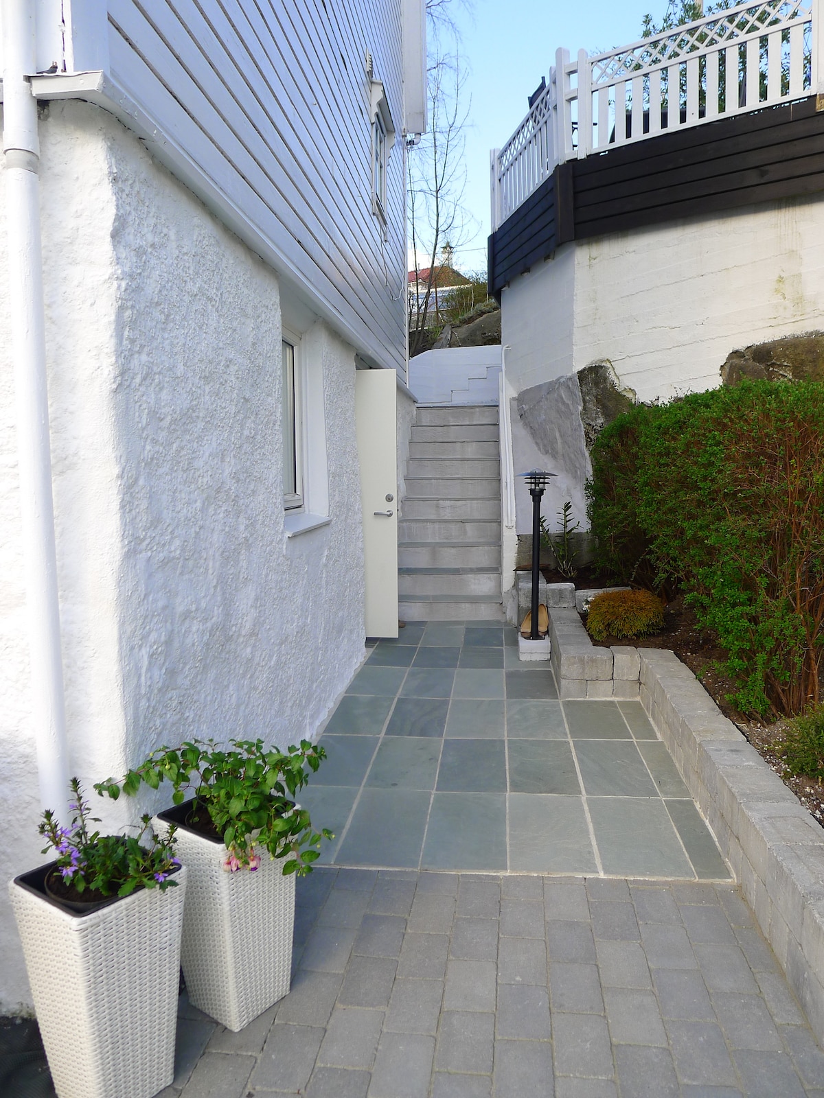 A pathway lined with slate tiles leads to a set of stairs, flanked by planters containing green plants. A white door is visible at the end of the path, while a well-maintained garden area with shrubs and a lantern is positioned to the right.